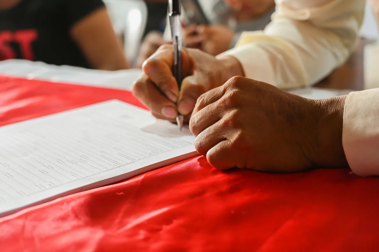 A close-up of hands signing a document with a pen on a red tablecloth. The background is blurred, suggesting a formal or official setting.