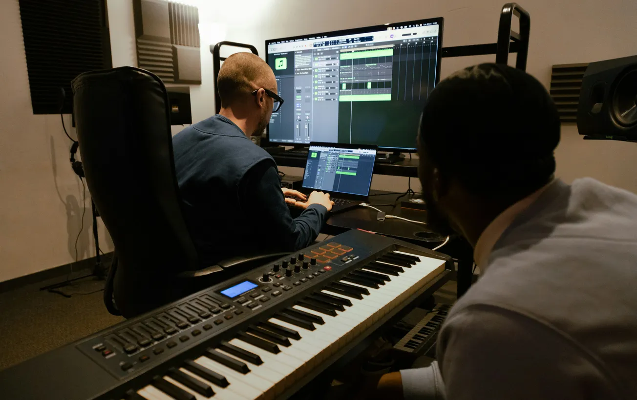 Two people in a music studio work on a digital audio workstation. One, wearing glasses, is at a desk with a computer, while the other observes. A keyboard is in the foreground.