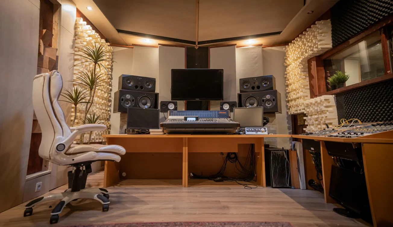 A cozy music studio with a mixing desk, multiple speakers, and a large monitor. A white ergonomic chair is on the left, surrounded by soundproofing panels.