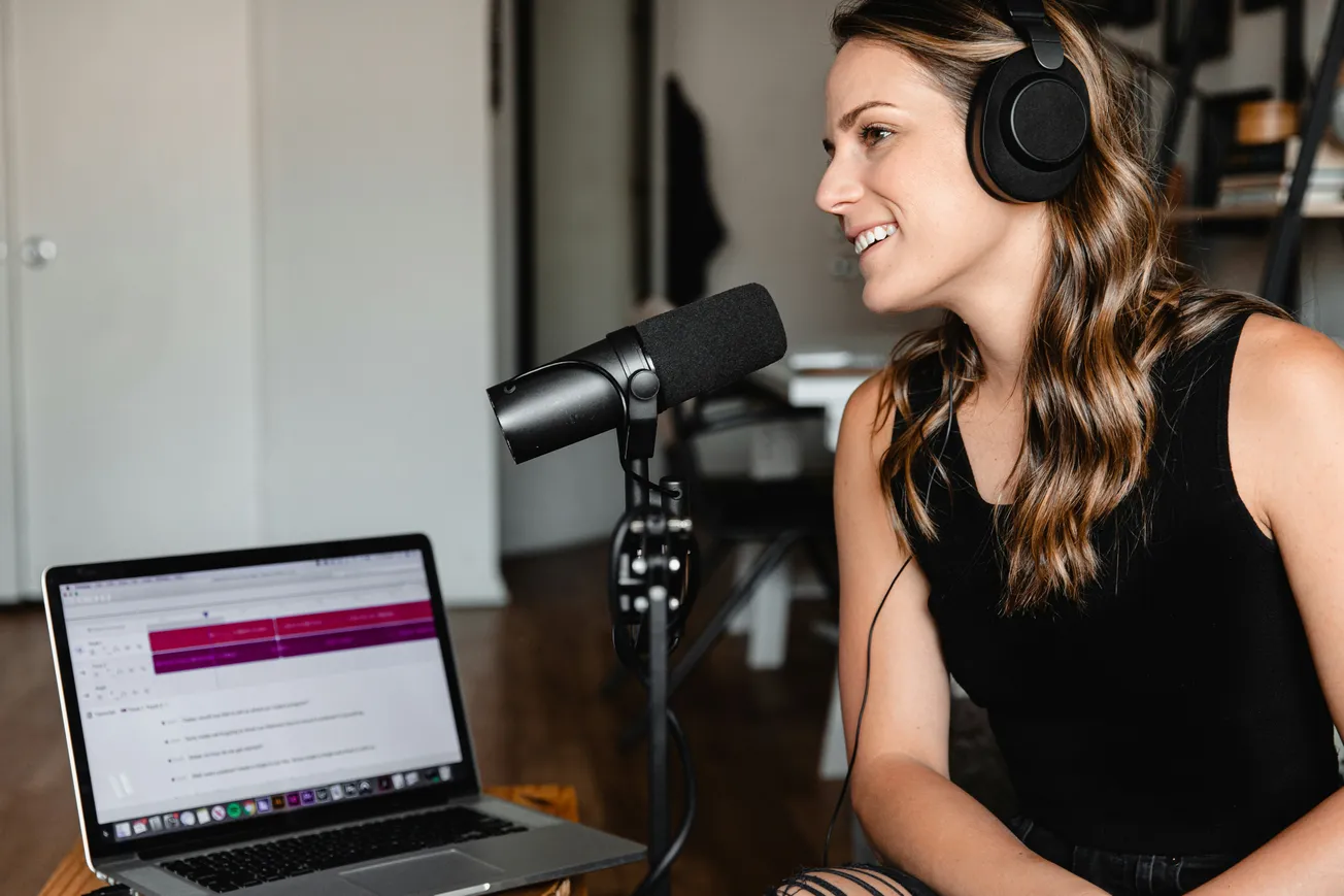A woman in headphones sits at a desk, speaking into a microphone. She smiles while looking at an open laptop, suggesting a podcast recording.