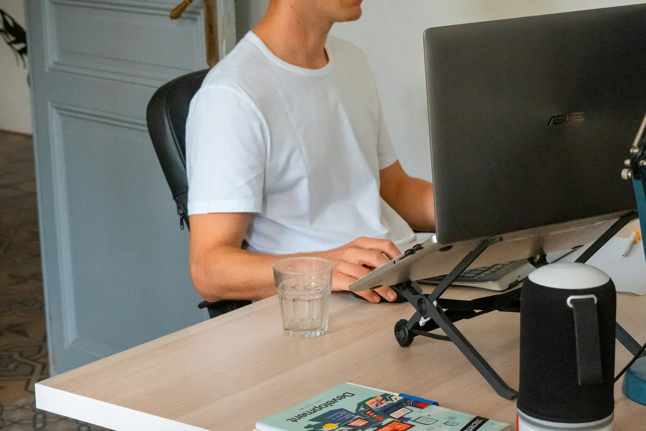 A person in a white shirt sits at a desk, typing on a laptop with a monitor. A glass of water and a book titled "Presentation" are on the desk.