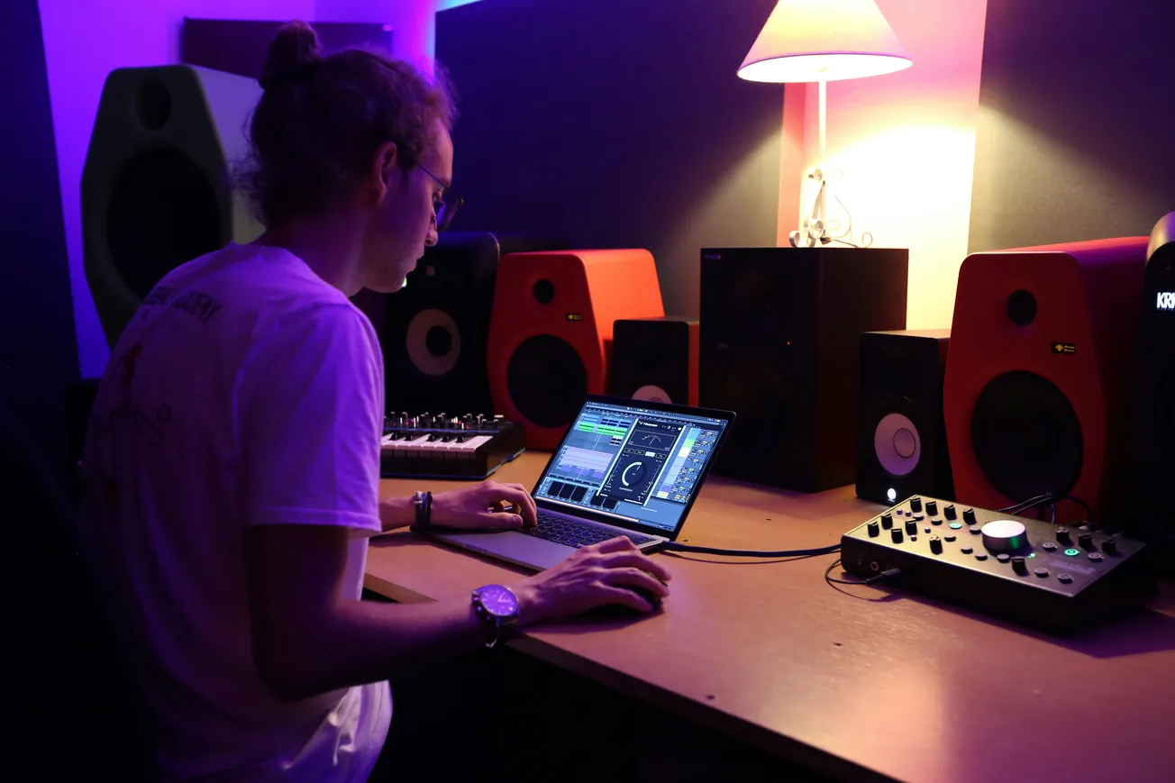 A person working on music editing software on a laptop, surrounded by speakers and sound equipment in a dimly lit studio with a relaxed ambiance.