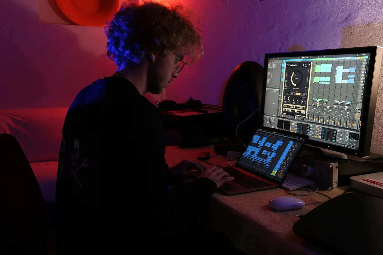 A person with curly hair works on music production at a dimly lit desk with two screens showing audio software. The ambiance is creative and focused.