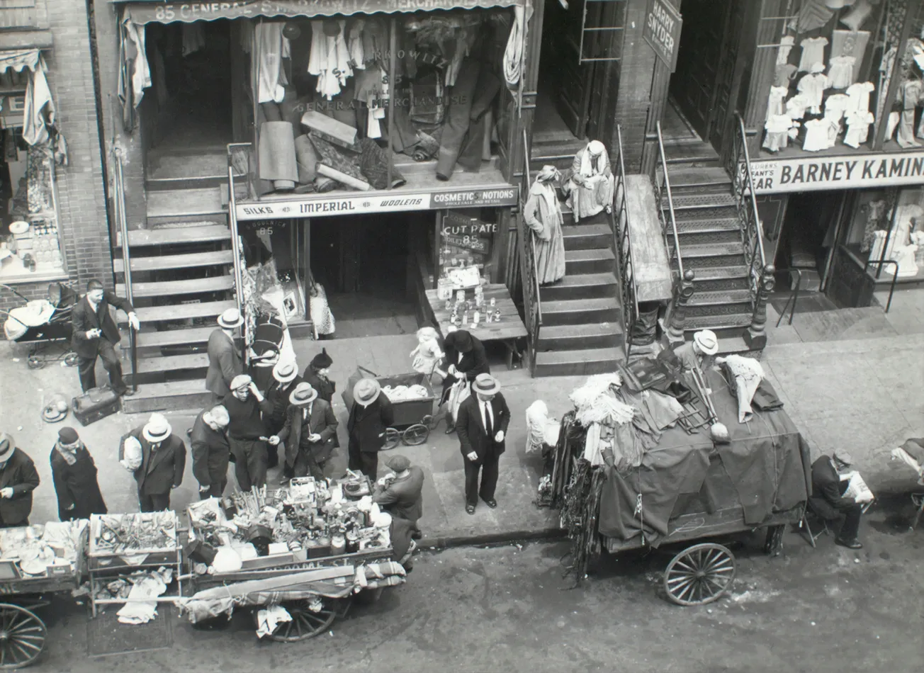 A bustling 1930s street market scene with vendors selling goods from carts, surrounded by formally dressed men in hats. Shops in the background display clothing.