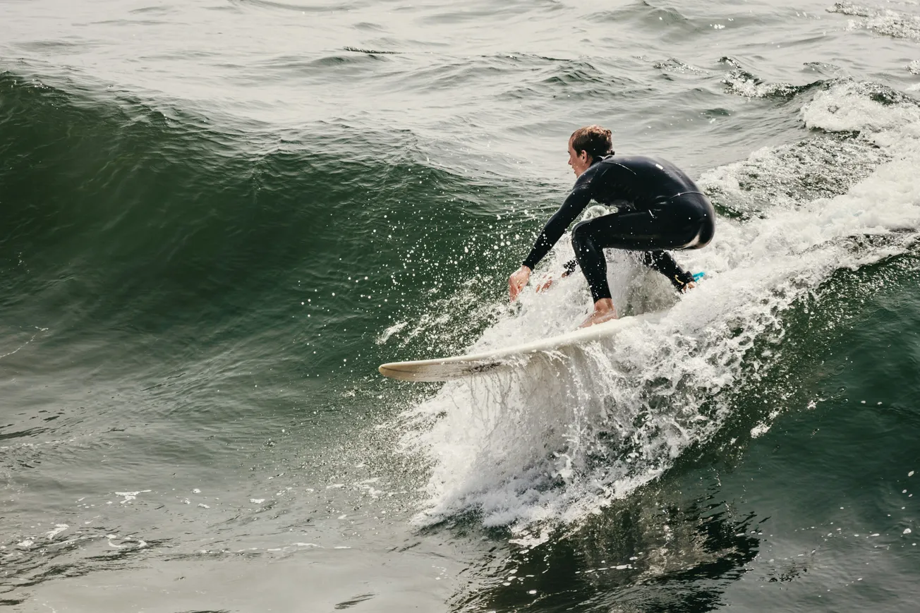 A surfer in a black wetsuit rides a large, curling wave on a white surfboard. The water splashes dynamically, conveying excitement and motion.