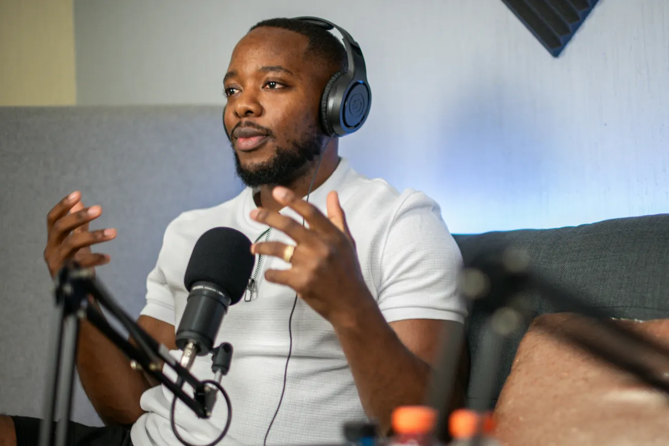 A man wearing headphones speaks passionately into a microphone, seated in a cozy studio with acoustic foam panels. He gestures expressively, conveying engagement.