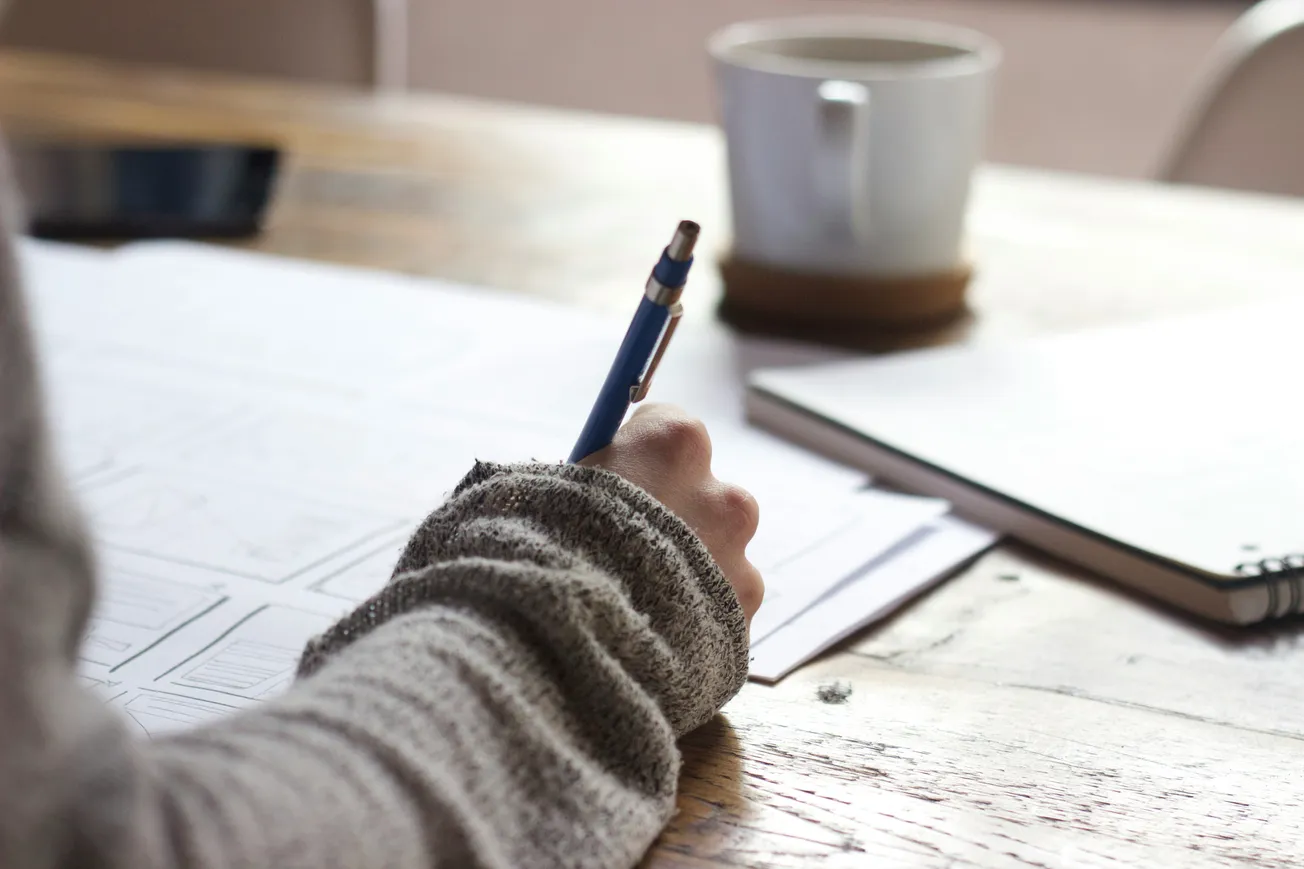 A person wearing a gray sweater writes with a pen on paper at a wooden table, with a white mug and notebook nearby, conveying a focused atmosphere.