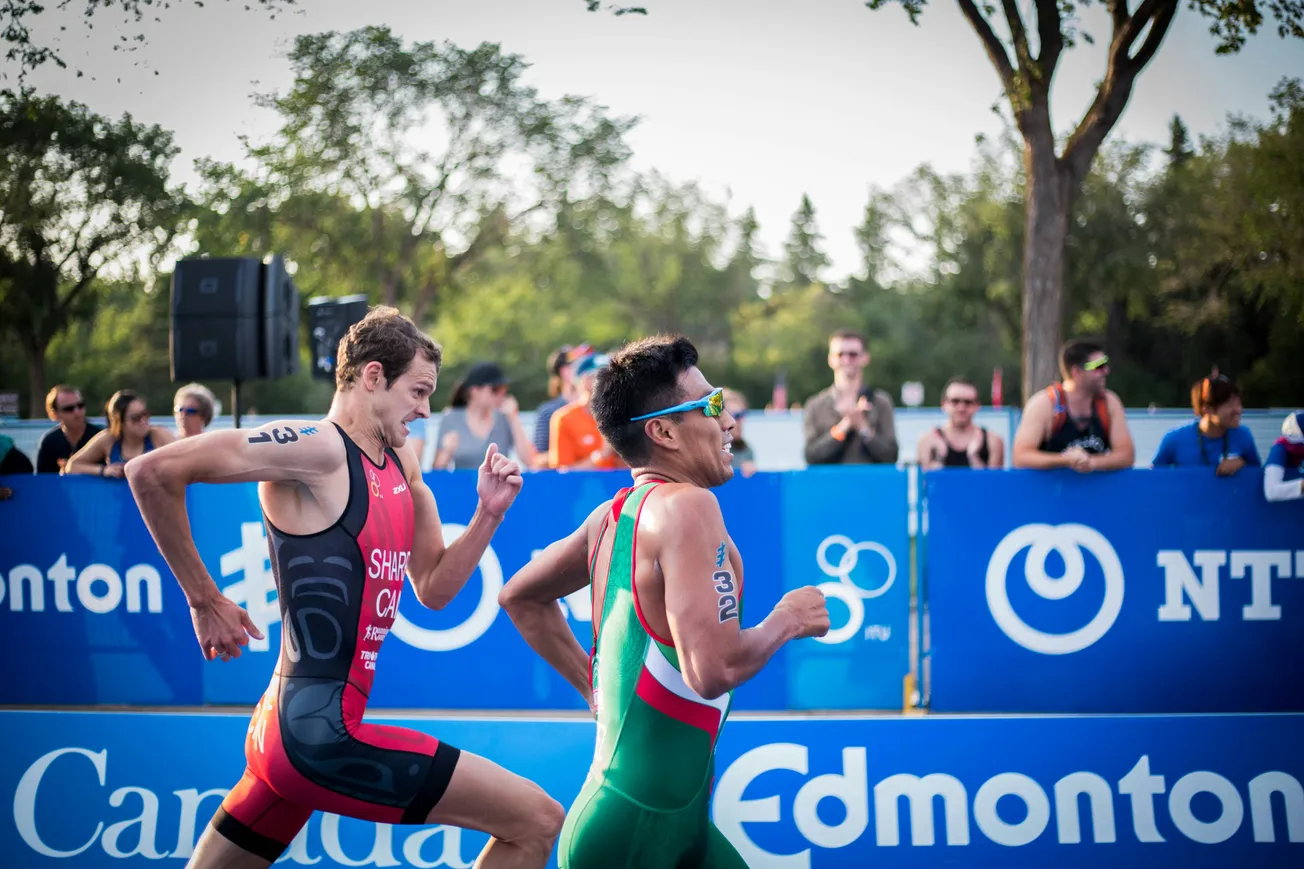 Two triathletes sprint side by side during a race, surrounded by cheering spectators. Trees and event signage are visible in the background.