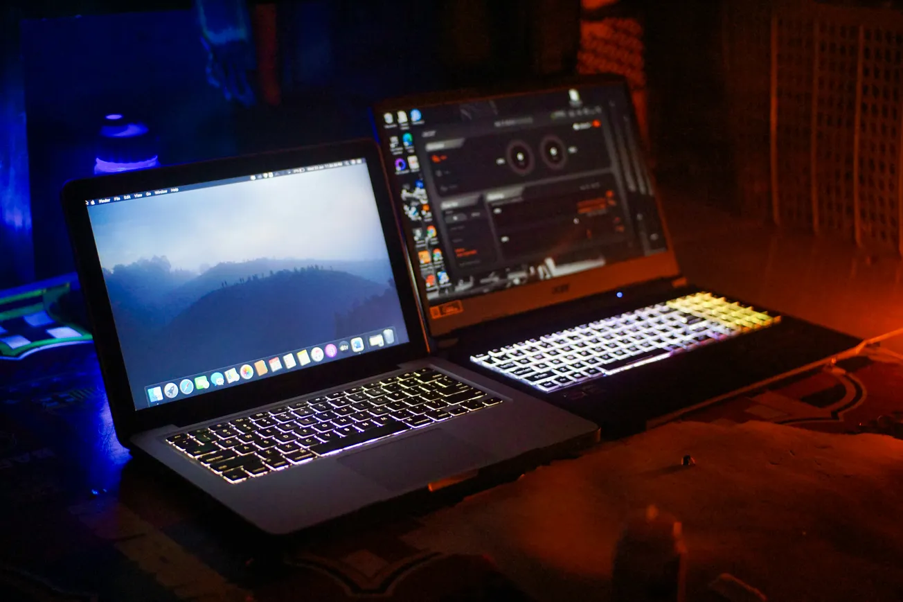 Two laptops with illuminated keyboards on a dimly lit desk. One displays a mountain scene wallpaper; the other shows a software interface. Cozy, tech-focused vibe.