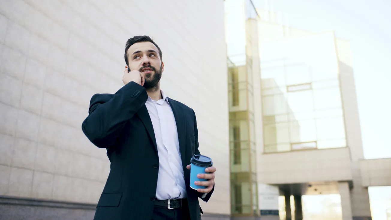 A man in a dark suit holds a blue coffee cup and talks on his phone outside a modern glass building. The scene conveys a professional, focused demeanor.