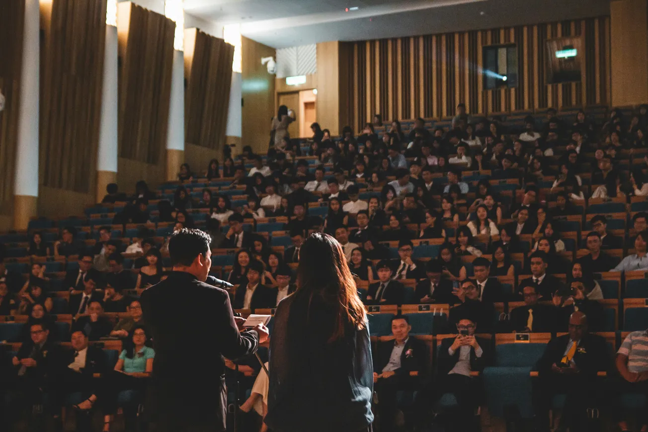 Two people address a large, seated audience in a dimly lit auditorium, creating an atmosphere of focused attention and engagement.