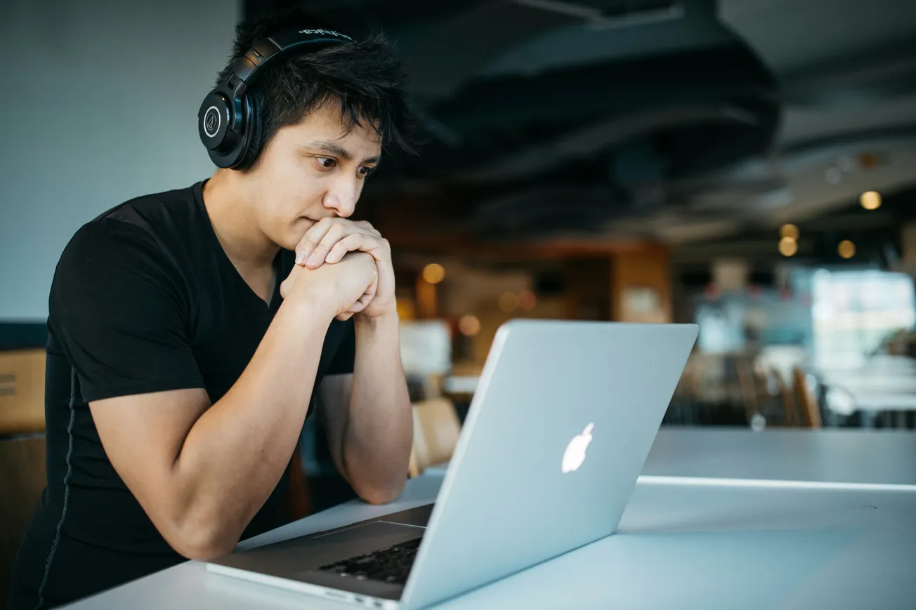 A man with headphones sits at a table, focused on his laptop while working or engaging in an online activity.