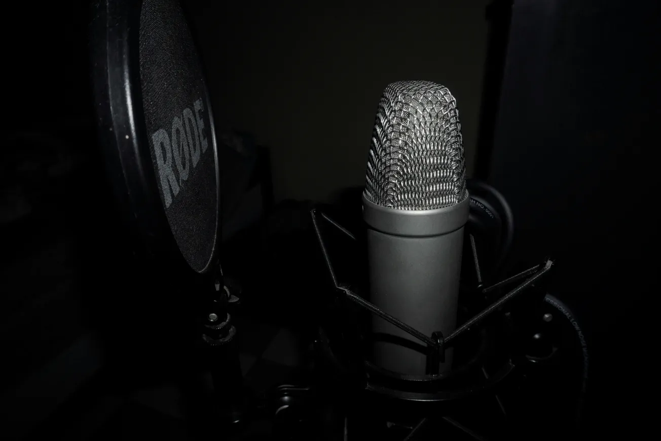 Close-up of a studio microphone with a pop filter, set against a dark background. The mic is silver with a textured grille.