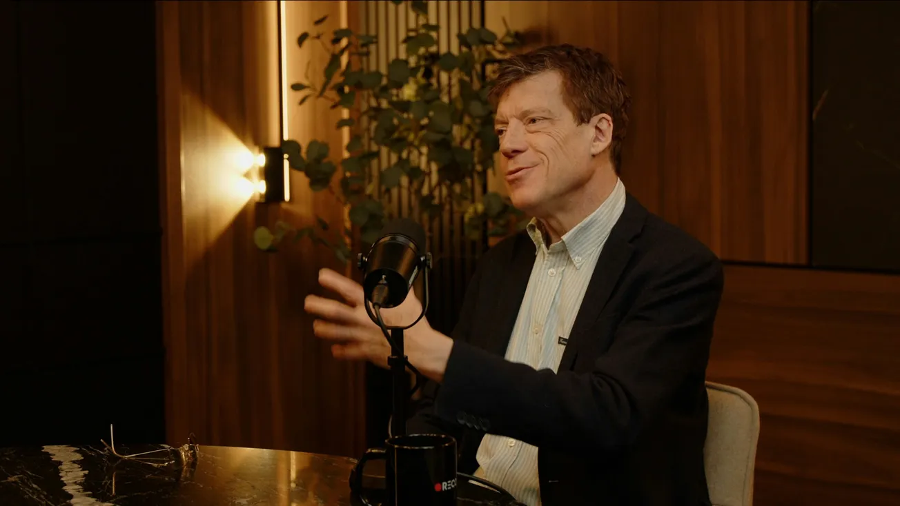Man in a dark blazer and striped shirt speaks into a microphone in a warmly lit room, gesturing with his hands. A mug and glasses are on the table.